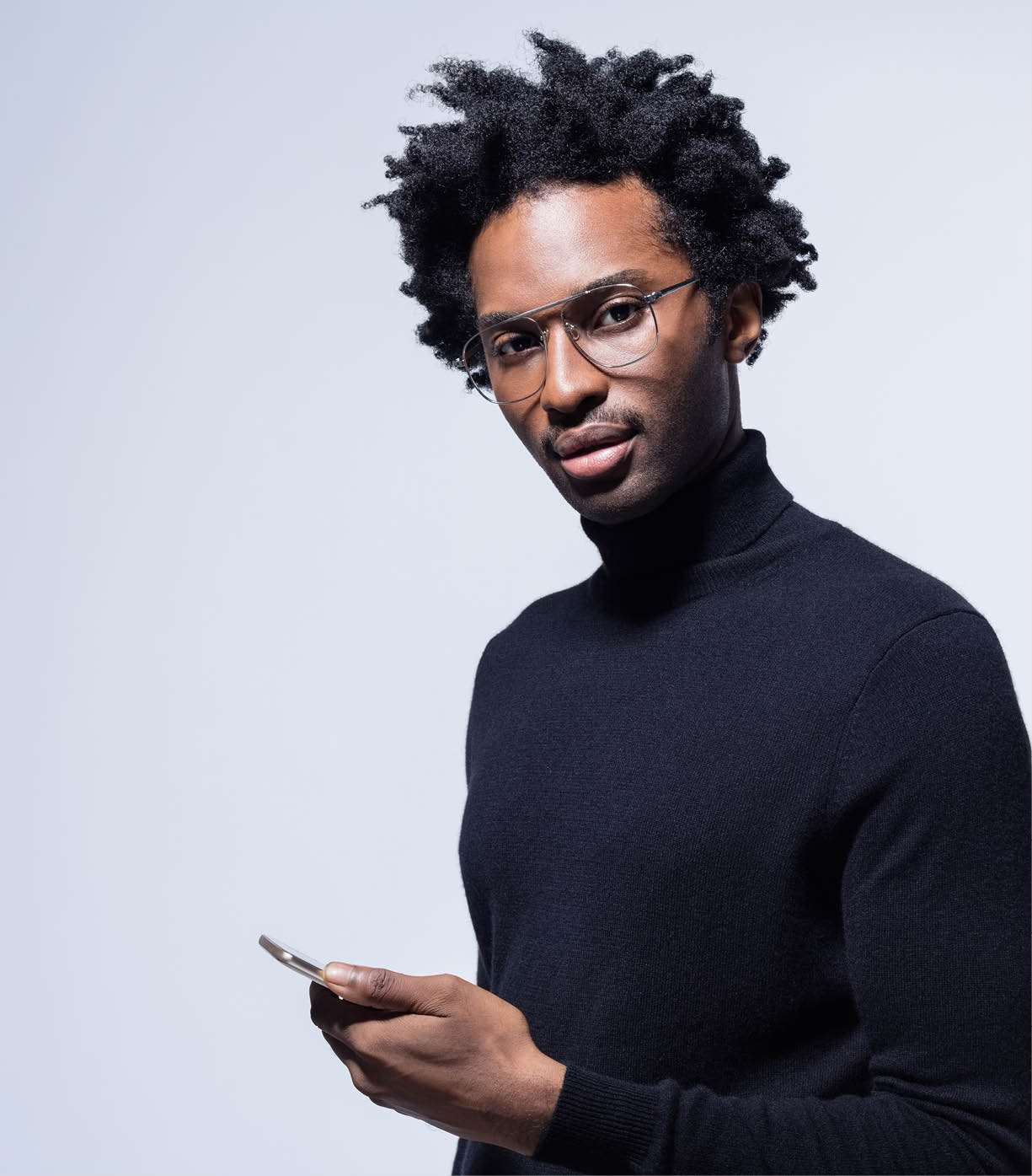 Afro american young man wearing black turtleneck and glasses holding smart phone in hand, looking at camera  Studio shot on grey background 